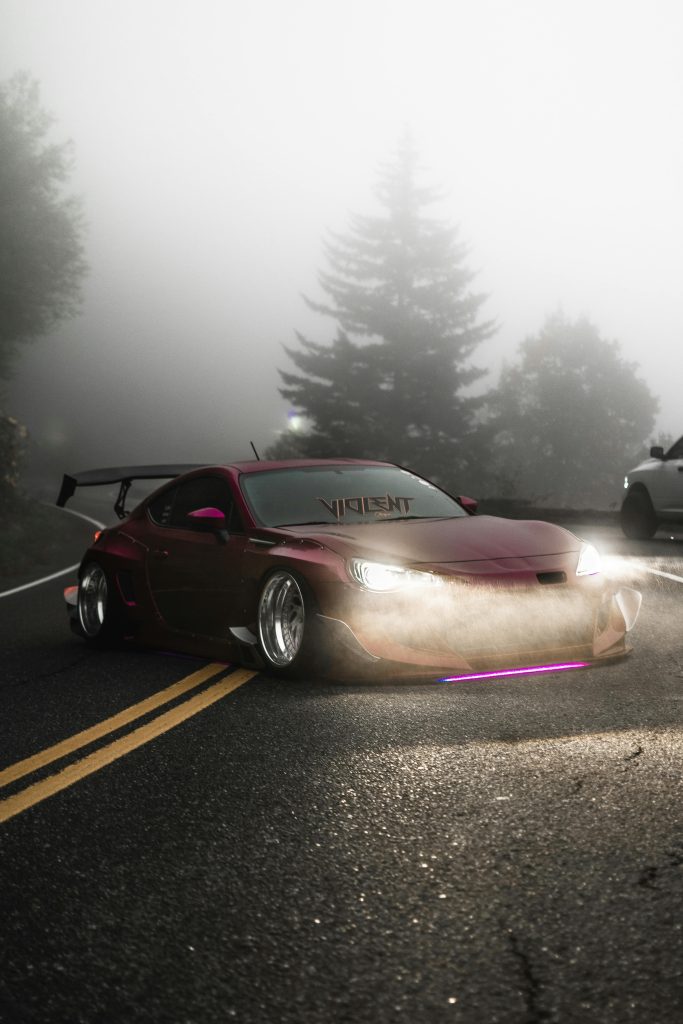 Red sports car with headlights on, driving through a foggy mountain road with pine trees.